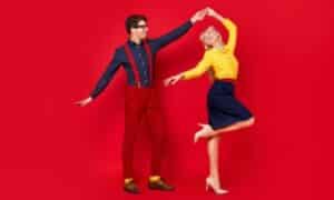 Una coppia balla su uno sfondo rosso. Lei ha una camicetta a amiche lunghe gialla e una gonna a tubino lunga fino al ginocchio blu scuro, lui ha i pantaloni e le bretelle rosse con una camicia blu