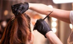 Foto in primo piano delle mani di un'esperta parrucchiera che con guanti neri e pennello passa la tinta sulla base laterale di una donna castana